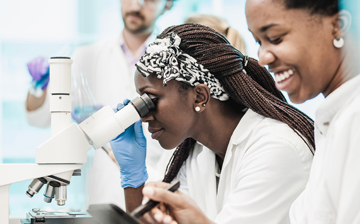 One woman looking in a microscope and another woman looking away and smiling.