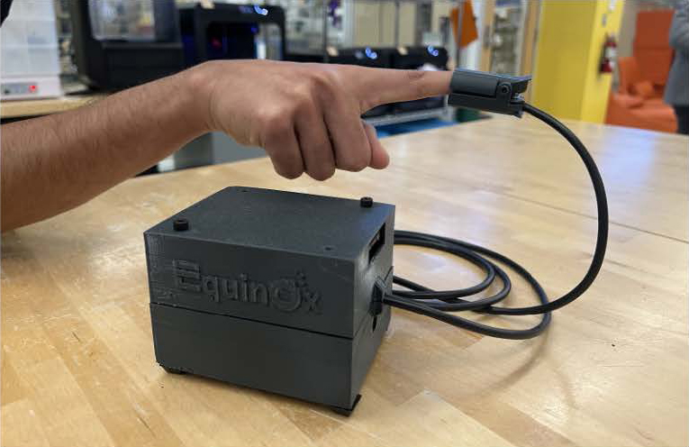 A photo a black box attached to a pulse oximeter attached to a finger.