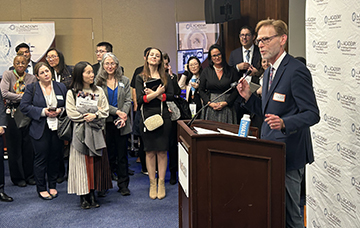 People stand around man speaking from a podium microphone