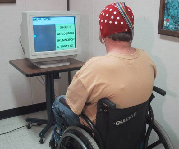Foto de un hombre en silla de ruedas sentado frente a una computadora con una gorra roja con electrodos EEG