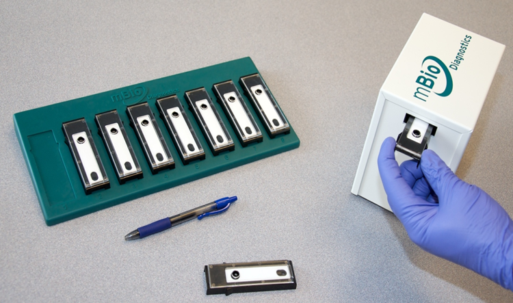 A photo of small chip devices on a desk next to a pen for scale and a purple gloved hand placing one of the devices into a box labeled mBio Diagnostics