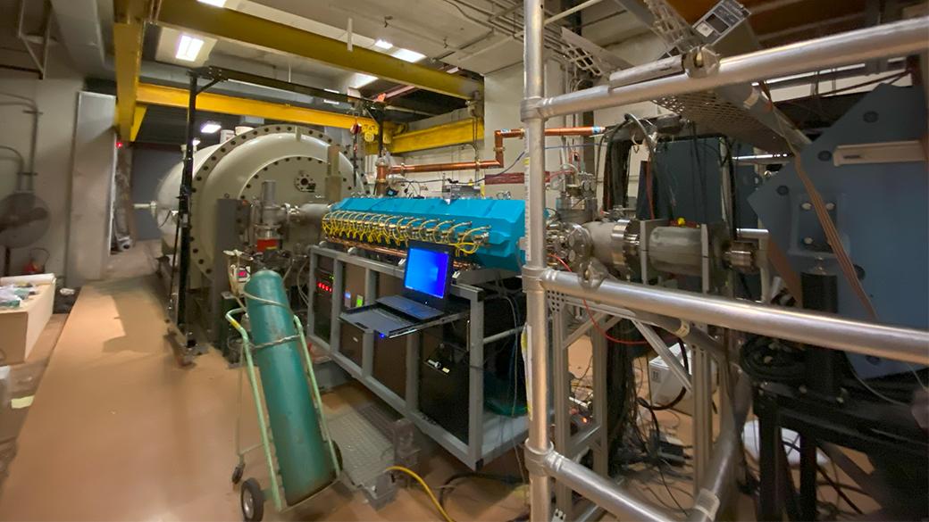 The photo shows the accelerator laboratory, a large room with complex machinery and equipment. In the center, there is a large cylindrical apparatus with multiple connected components, including a blue section with numerous valves and pipes. A laptop with a blue screen is placed on a cart next to the machinery. The space has overhead lighting and various structural elements, such as pipes and scaffolding.