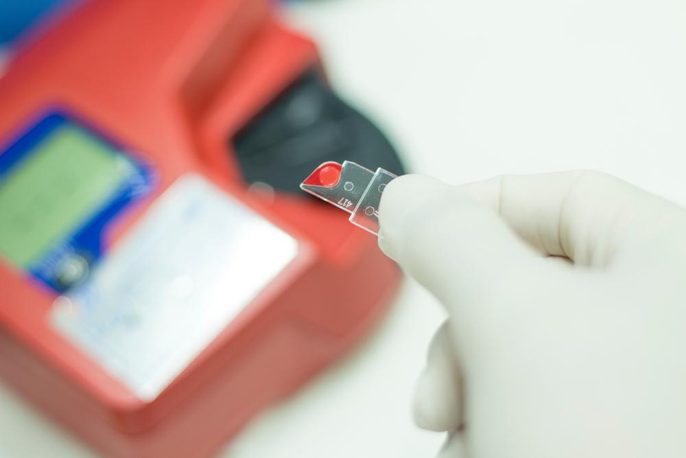 in this image a gloved hand holds a small glass slide with a drop of red liquid, possibly blood, on it. In the background, a red laboratory device with a digital display is visible but out of focus.