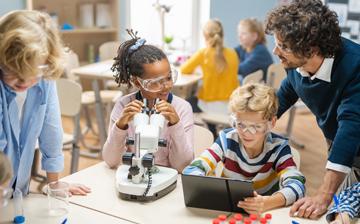 Three children and a teacher with one child looking in a microscope and another looking at a tablet